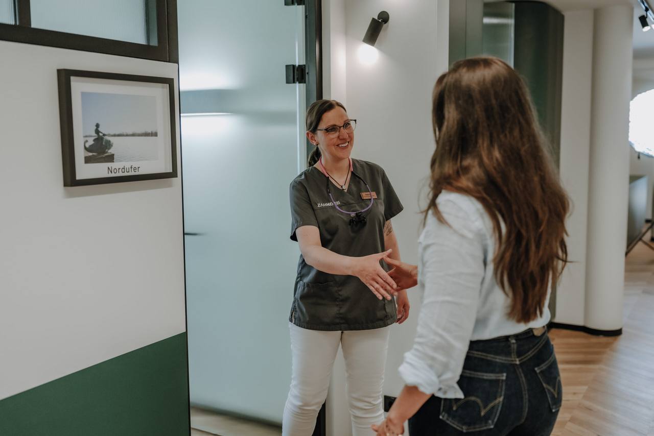 Mitarbeiterin begrüßt eine Patientin per Handschlag vor dem Behandlungszimmer Nordufer