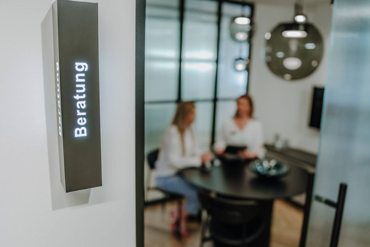 Beleuchtetes Beratungs-Schild, im Hintergrund ein Beratungsgespräch am runden Tisch