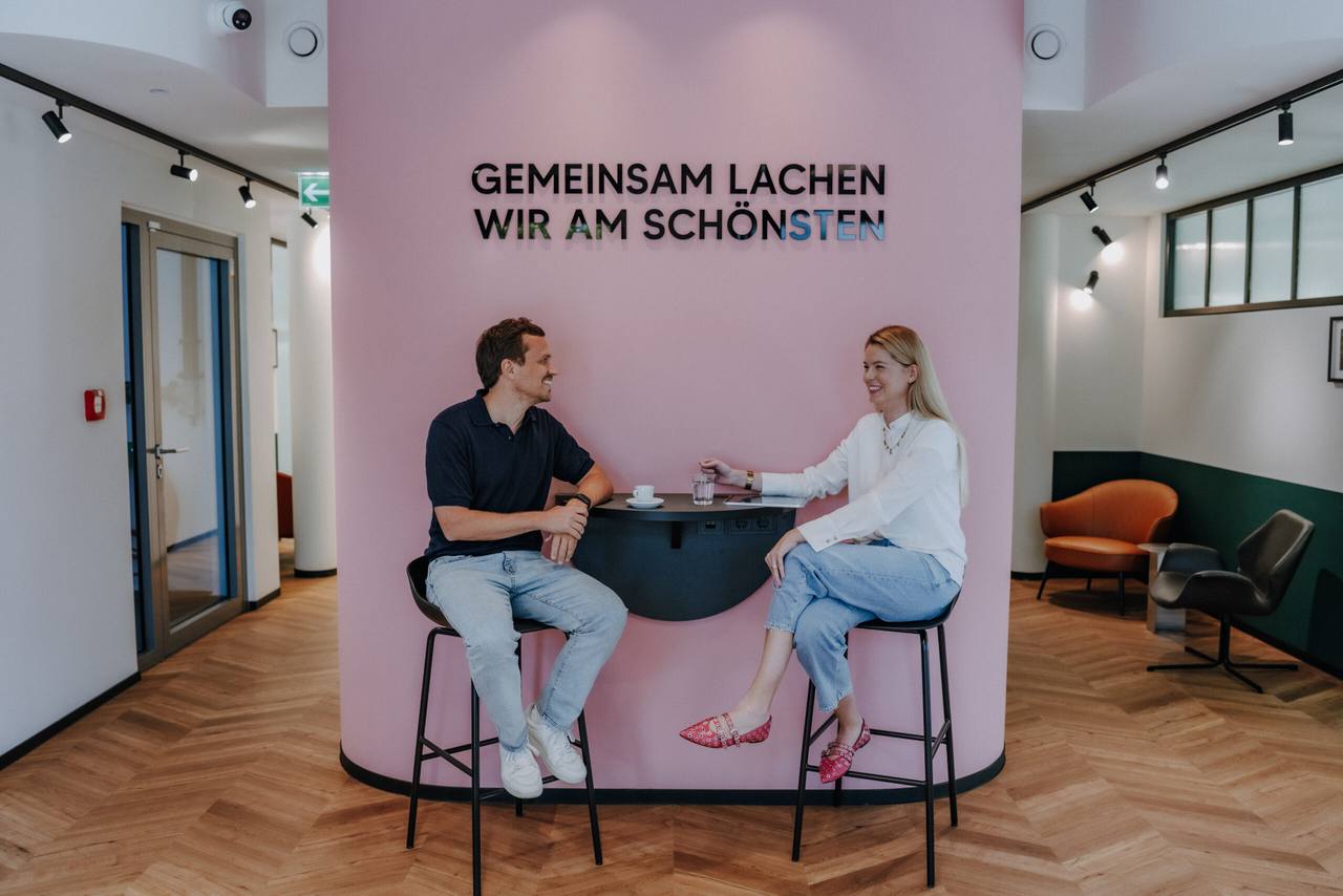 Zwei Menschen im Gespräch am Stehtisch vor der rosa Wand mit dem Schriftzug GEMEINSAM LACHEN WIR AM SCHÖNSTEN