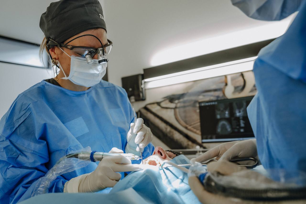 Chirurgin mit Haube und Lupenbrille während eines oralchirurgischen Eingriffs, im Hintergrund die DVT-Aufnahme am Monitor
