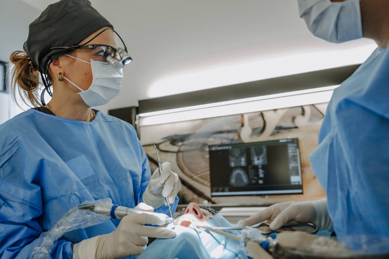 Chirurgin mit Lupenbrille und Assistenz während eines oralchirurgischen Eingriffs, im Hintergrund die 3D-Röntgenplanung