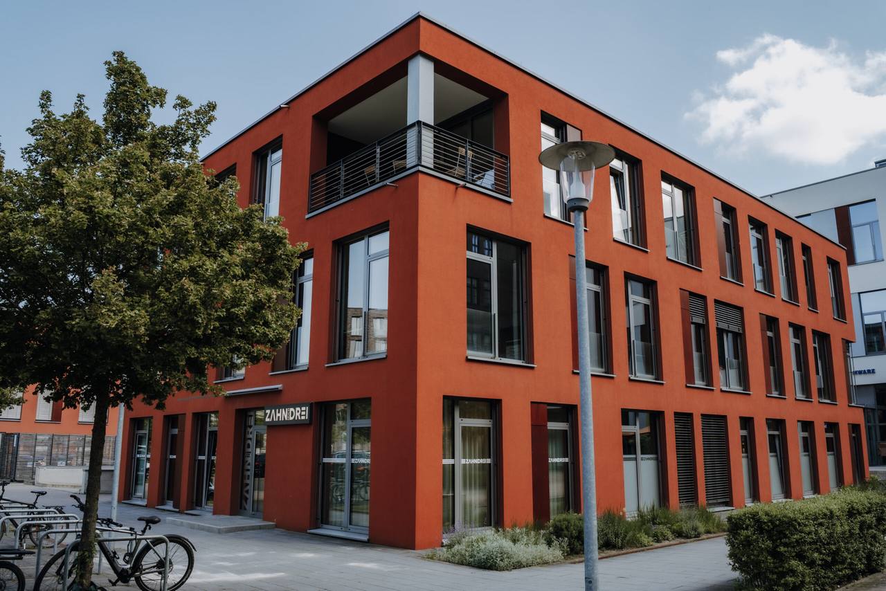 Das rote Praxisgebäude von zahnhochdrei auf dem Hanomaggelände mit ZAHNHOCHDREI-Schriftzug, Baum und Fahrradständern